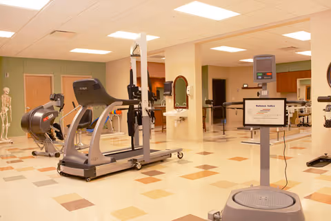 A spacious physical therapy or rehabilitation room with exercise equipment including a treadmill, stationary bike, and balance testing machine. The room has a tiled floor with a pattern of beige and brown squares, light green walls, and bright overhead lighting. A skeleton model is visible in the background near a door.
