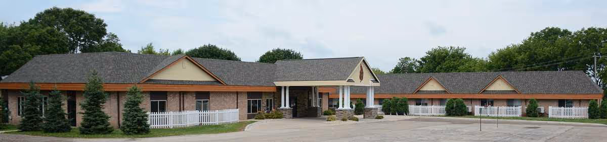 Wide exterior view of a single-story senior living facility building with a pitched roof, brick walls, white picket fences, and a covered entrance supported by white columns. The building is surrounded by trees and a paved driveway.
