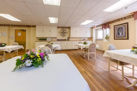 Dining room with several tables draped in white tablecloths and floral centerpieces, upholstered chairs, wood flooring, and cabinetry along the back wall.