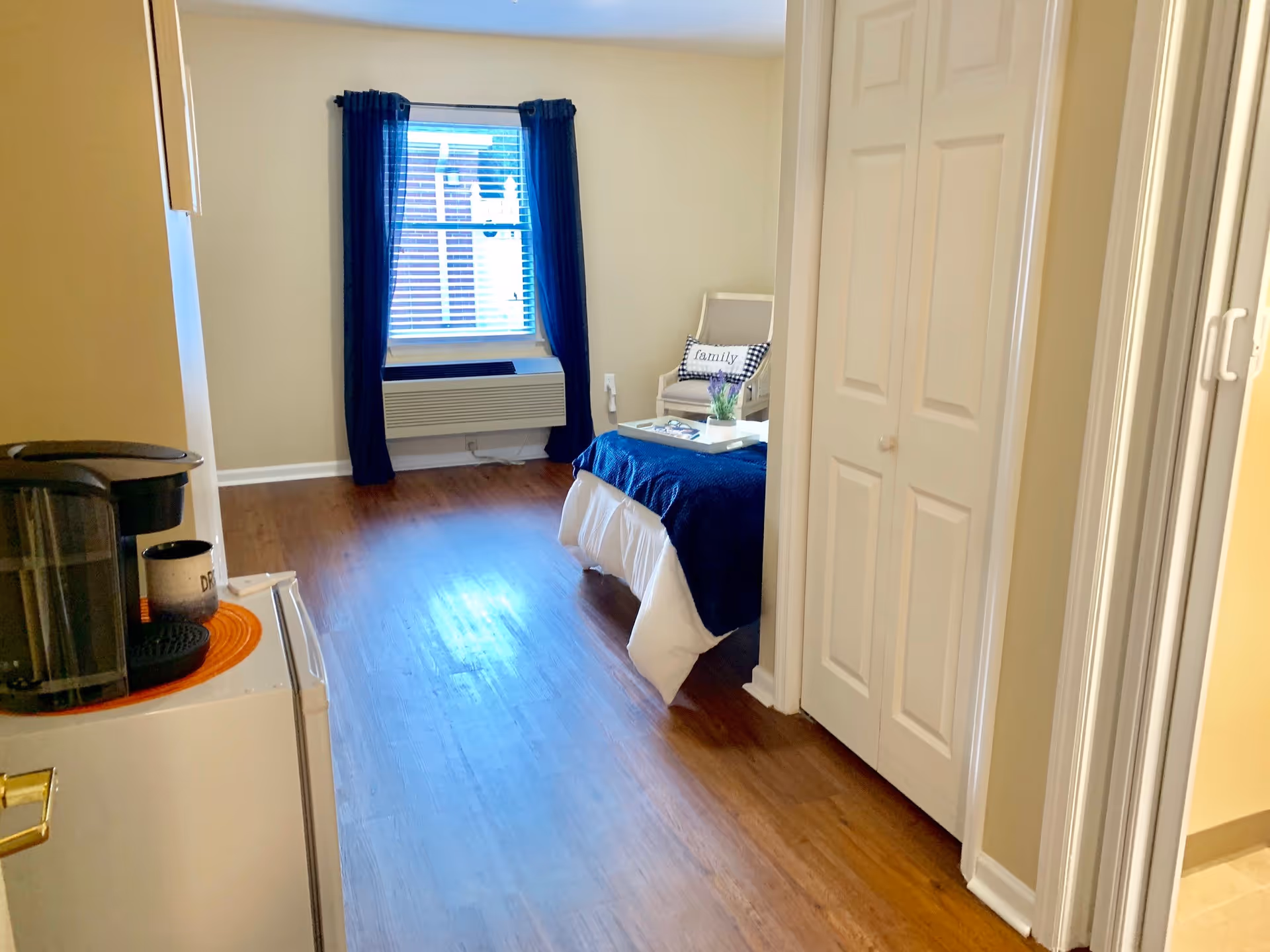 View of a senior living facility room with wooden flooring, a bed with a blue blanket, a small armchair with a pillow that says 'family', a window with blue curtains, and a small kitchenette area with a coffee maker and a mug.