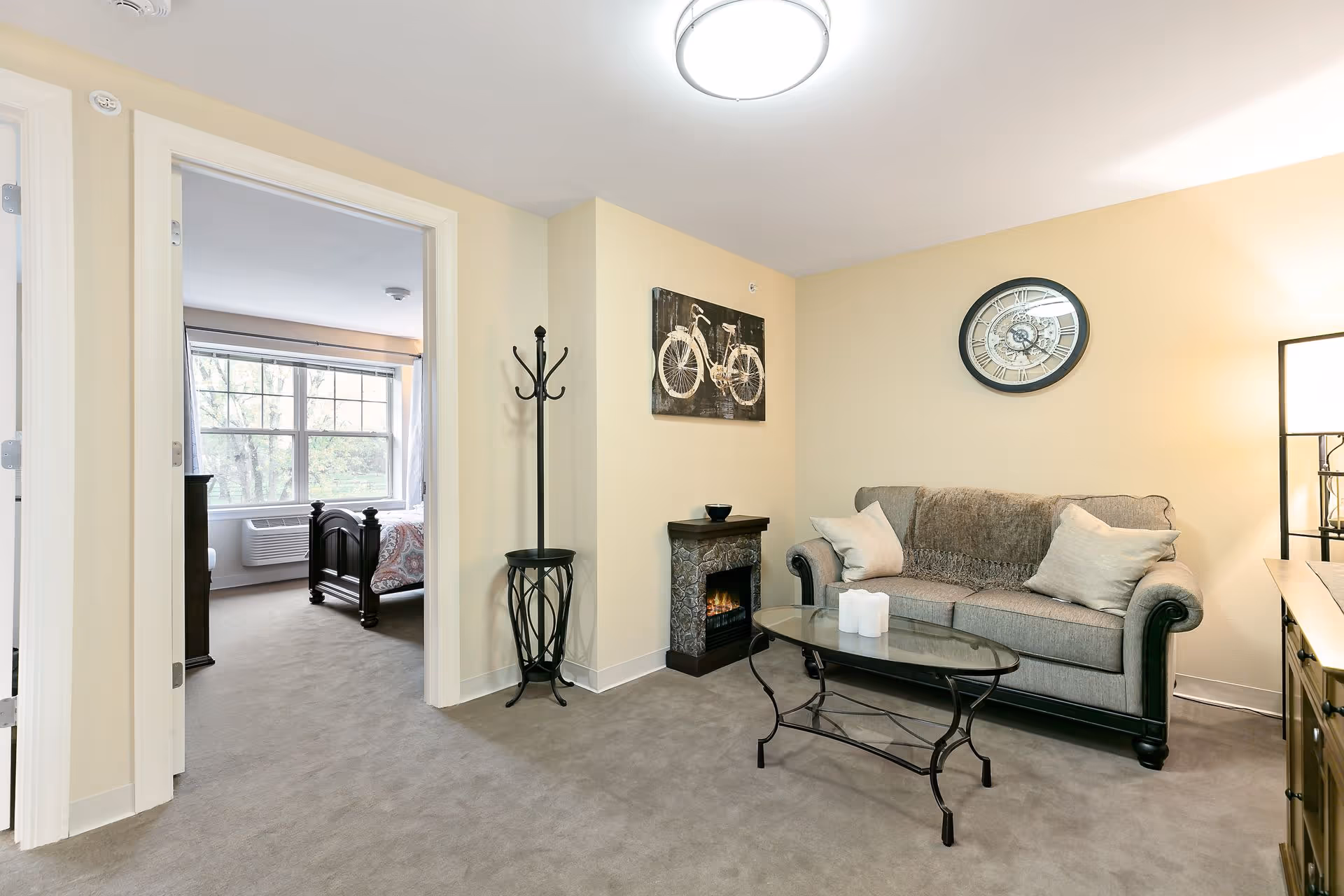 A cozy living room with a gray sofa adorned with two pillows and a throw blanket, a glass coffee table with candles, a small electric fireplace, a coat rack, and wall decorations including a bicycle painting and a round clock. An adjacent bedroom with a large window and a dark wooden bed frame is visible through an open doorway.
