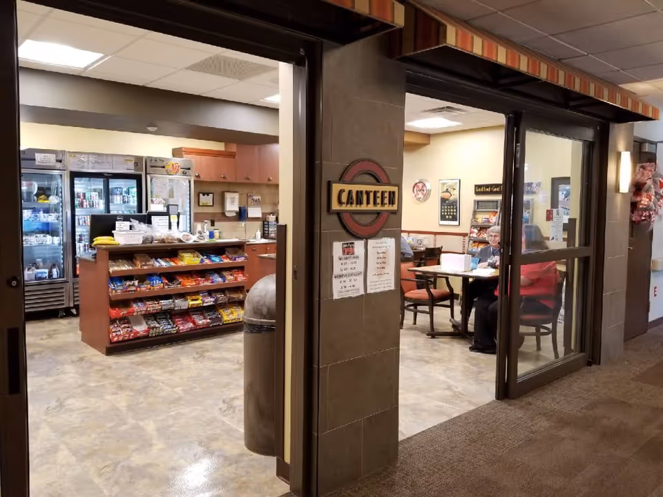 Interior view of a canteen area in a senior living facility. The canteen has shelves stocked with snacks and refrigerated coolers with beverages. There are tables and chairs where elderly residents are seated. The entrance has a sign that reads 'CANTEEN'.