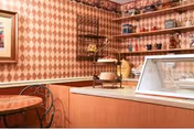 Interior view of a small cafe or snack bar area with a round wooden table and metal chairs, a counter with a glass display case, shelves with various jars and containers, and patterned wallpaper with a diamond design.