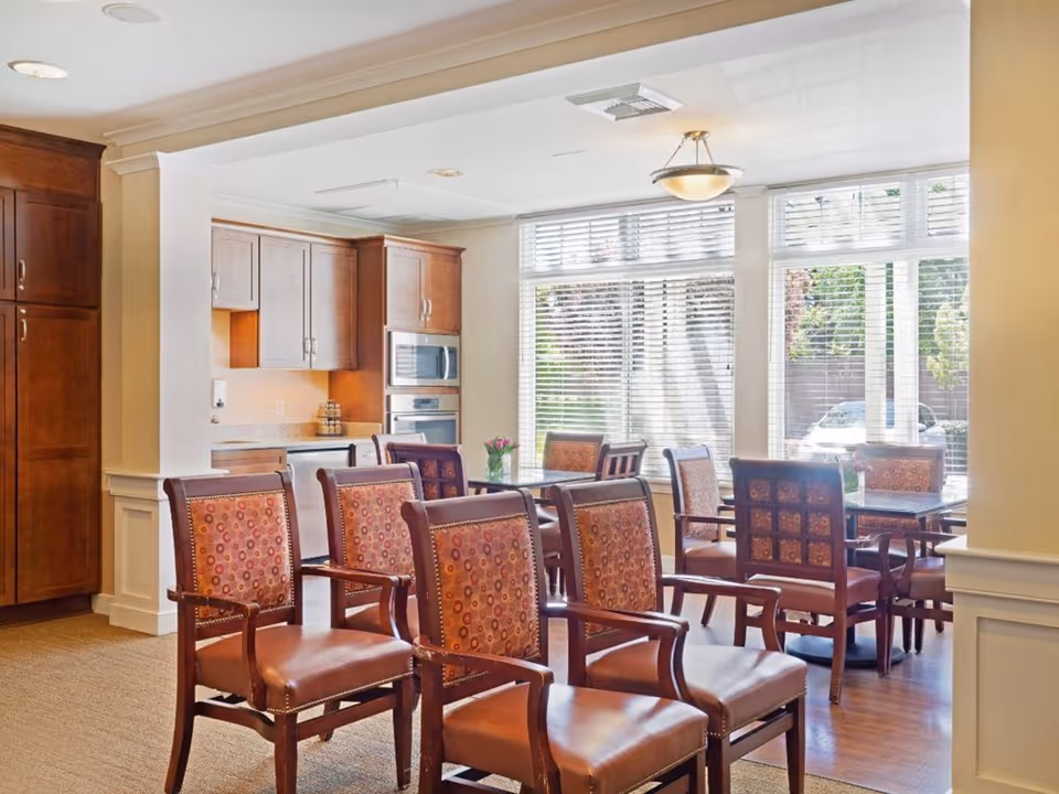 Bright dining area with wooden chairs and tables in front of a kitchenette and large windows with blinds.