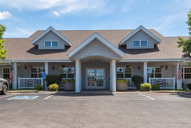 Front exterior view of a single-story assisted living facility building with a peaked roof, two dormer windows, a covered entrance supported by columns, American flags, and handicap parking spaces in front.