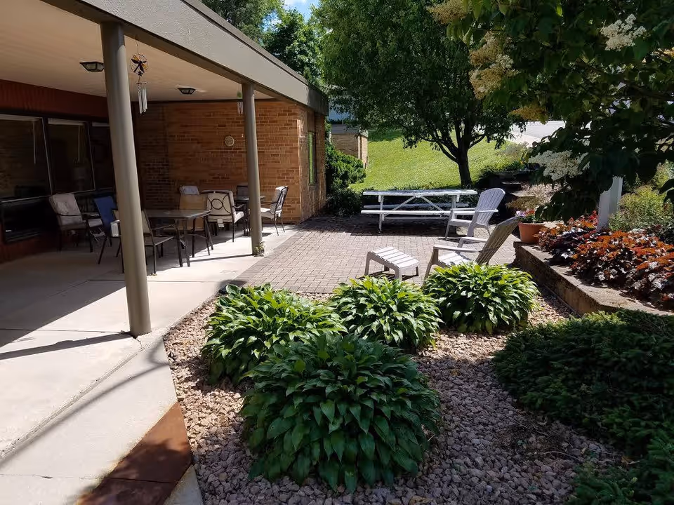 Outdoor patio area at Traditions of Preston with a covered seating area featuring tables and chairs, a paved section with Adirondack chairs and a small footrest, surrounded by green bushes, plants, and trees under a sunny sky.