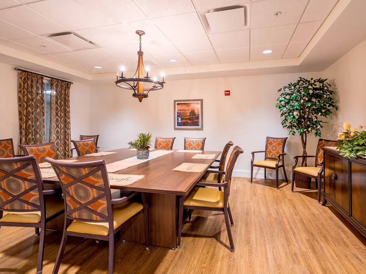 A well-lit dining room with a large wooden table surrounded by ten chairs with patterned backs and yellow cushions. The room features a hanging chandelier, a framed picture on the wall, patterned curtains on the window, a wooden sideboard with plants and candles, and a tall potted plant in the corner. The floor is wooden, and the walls are painted light beige.