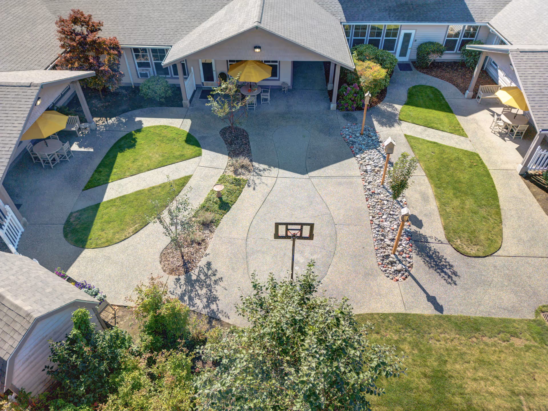 Aerial view of a courtyard with winding walkways, grassy islands, seating areas with yellow umbrellas, landscaping and a small basketball hoop.