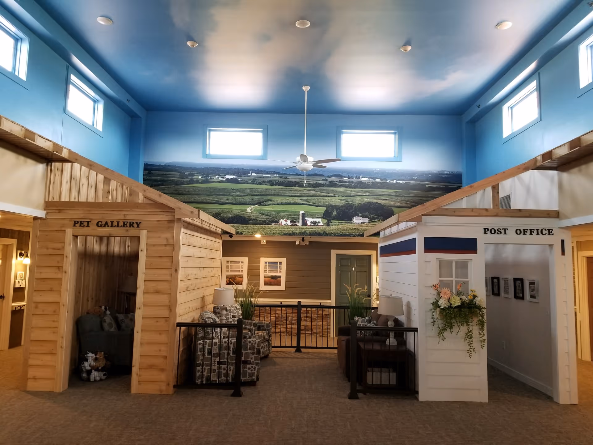 Interior of a senior living facility with a high ceiling painted to resemble a sky with clouds. The room features two small wooden structures labeled 'PET GALLERY' and 'POST OFFICE'. The pet gallery has a cozy seating area with a chair and pet toys, while the post office has a flower arrangement and framed pictures on the wall inside. There are several windows near the ceiling letting in natural light, and a large mural of a rural landscape with fields and farm buildings on the back wall.