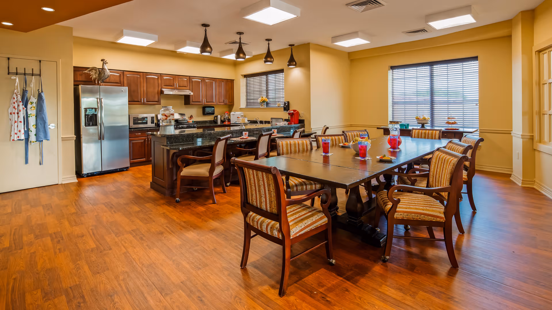 A spacious dining area with a large wooden table surrounded by eight cushioned chairs with striped upholstery. The table has several glasses filled with red liquid and some plates with snacks. In the background, there is a kitchen area with wooden cabinets, a stainless steel refrigerator, a microwave, and a kitchen island with additional seating. The room has wooden flooring, beige walls, and windows with blinds allowing natural light to enter.