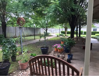 Shaded outdoor courtyard with wooden benches, potted plants, a paved walkway, and trees near a wooden fence.