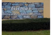 Sign on a stone wall that reads 'Gilmer Nursing & Rehabilitation Center' with a hedge and grass in front of it.