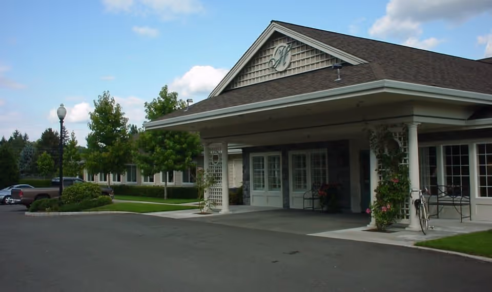 Exterior view of The Hampton senior living facility showing the front entrance with a covered drop-off area, columns, and a landscaped driveway with trees and parked cars under a partly cloudy sky.