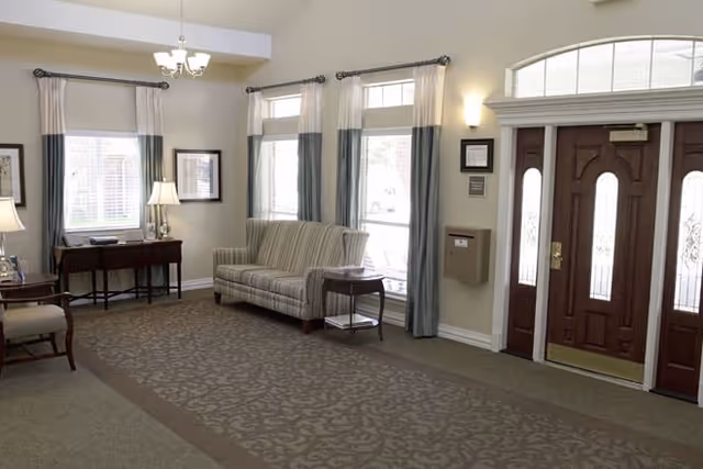 A well-lit living room area with a patterned carpet, a striped sofa, a small side table, a wooden desk with a lamp, and large windows with curtains. There is a wooden front door with glass panels and a chandelier hanging from the ceiling.