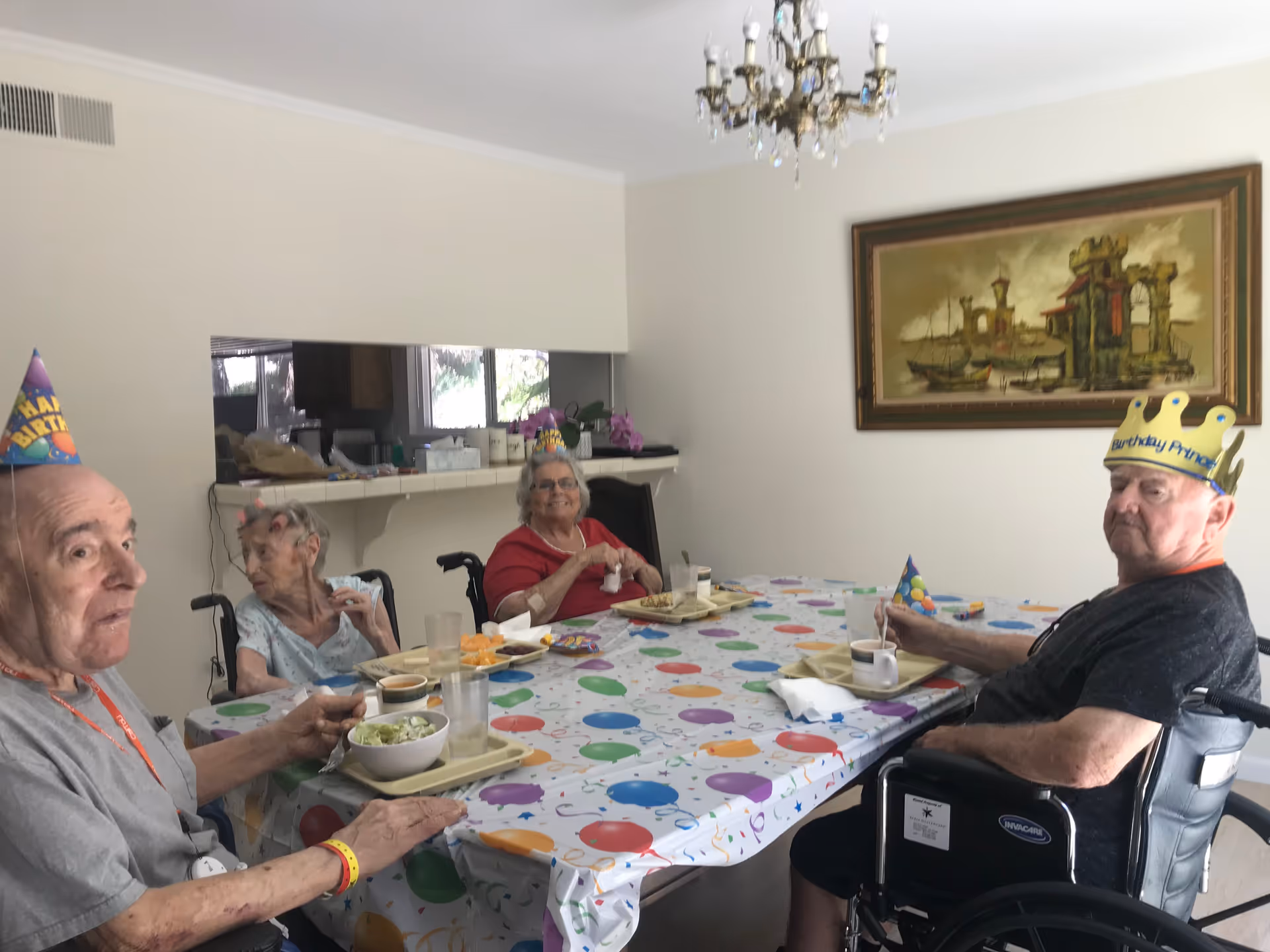 Four elderly people sitting around a dining table covered with a colorful balloon-themed tablecloth. They are wearing birthday hats and appear to be celebrating. The room has a chandelier, a large framed painting on the wall, and a pass-through window to the kitchen area.