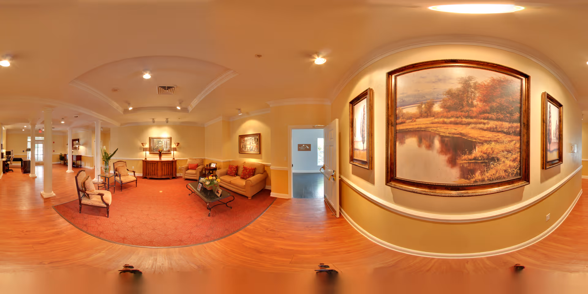 A spacious and warmly lit living room area in a senior living facility with wooden floors, beige walls, and white columns. The room features a seating area with a beige sofa, two armchairs, a glass coffee table with a flower arrangement, and framed landscape paintings on the walls. There is a red patterned rug under the seating area and a wooden sideboard with lamps and decorative items. An open door leads to another room with natural light.