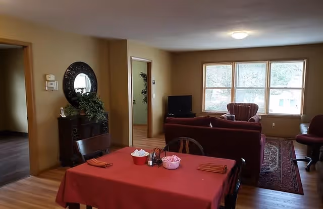 Interior view of a senior living facility showing a dining area with a table covered in a red tablecloth and set with napkins and condiments. Beyond the dining area is a living room with a red sofa, armchair, a TV on a stand, and a large window letting in natural light. There is a decorative mirror and plant on a small cabinet against the wall.