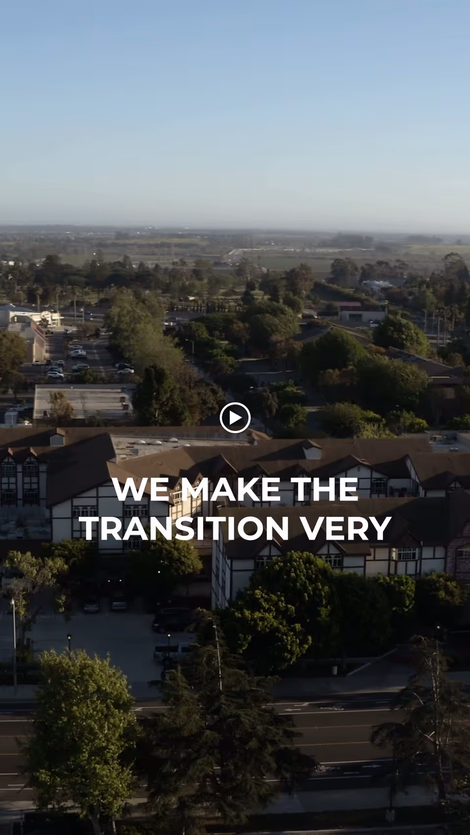 Aerial view of a large assisted living facility surrounded by trees and greenery with a road in the foreground and a clear sky above. The building has a Tudor-style architecture with multiple peaked roofs.
