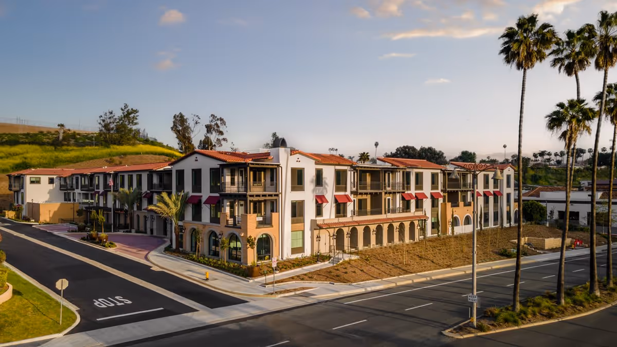 Exterior view of a multi-story senior living facility building with white walls, red tile roof, balconies, and red awnings. The building is situated at a street corner with palm trees lining the road and a clear sky above.