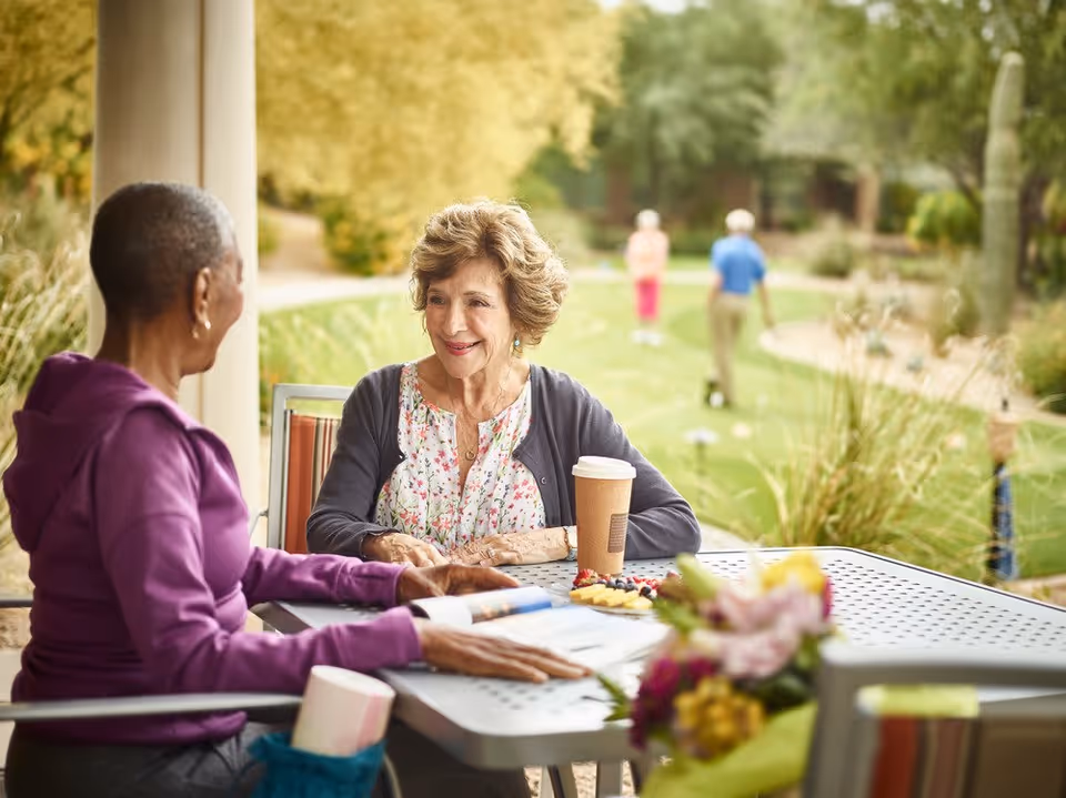 Two elderly women sitting at an outdoor table engaged in conversation, with a coffee cup and snacks on the table. In the background, two people are walking on a green lawn surrounded by trees and plants.