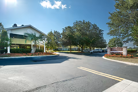 Exterior view of a senior living facility with a paved driveway, landscaped greenery including palm trees and large oak trees, and a sign that reads 'Emeritus at Canopy Oaks'. Several parked cars are visible near the building under a clear blue sky.