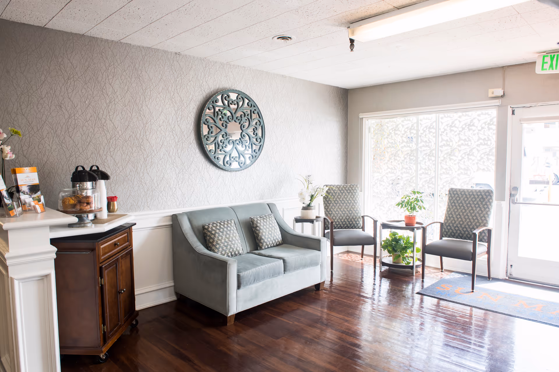 A bright and welcoming waiting area with a light blue loveseat with two patterned cushions, two armchairs with patterned upholstery, a small round table with potted plants, a wooden cabinet with refreshments including coffee dispensers and snacks, and a decorative round wall mirror. The floor is polished dark wood, and there is a glass door letting in natural light.