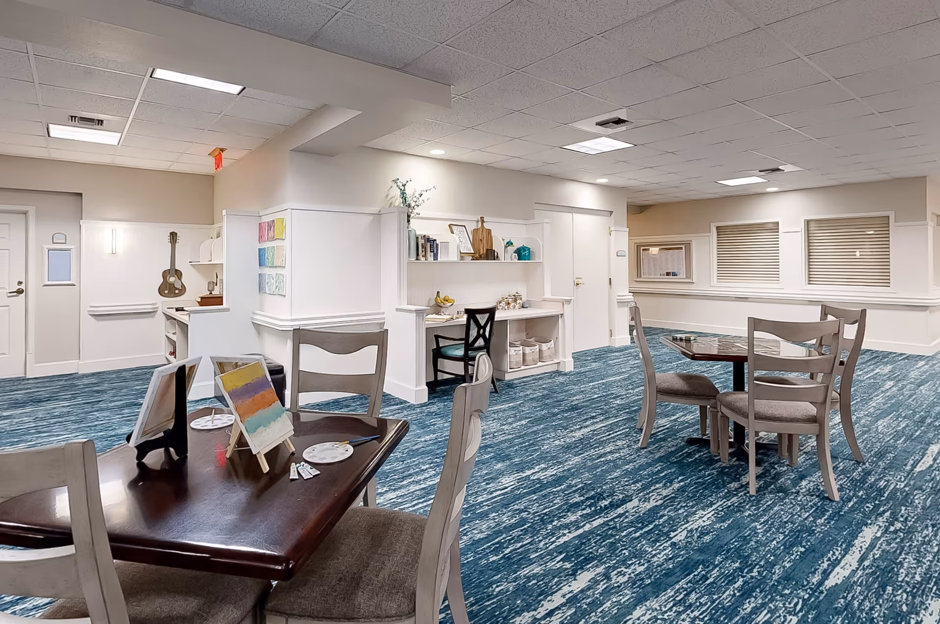 Interior room with blue patterned carpet, several wooden tables and chairs, and a small art station with paintings and art supplies. The walls are light-colored with shelves holding decorative items and books. There is a guitar hanging on the wall near a door.