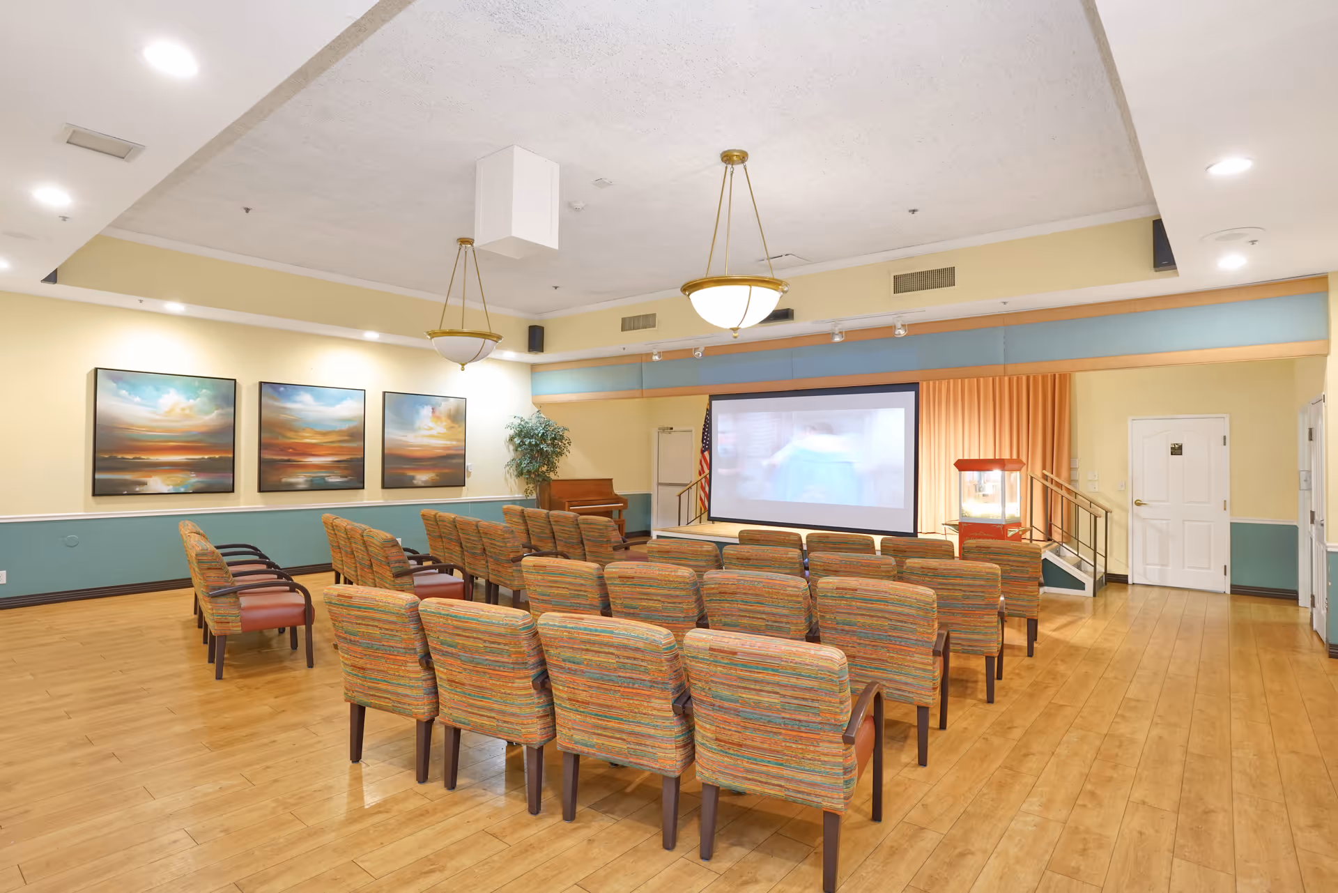 A community room with rows of colorful upholstered chairs facing a large projection screen. The room has wooden flooring, three landscape paintings on the left wall, two hanging ceiling lights, a piano in the corner, and an American flag next to the screen. There is also a popcorn machine near the screen and a door on the right side.