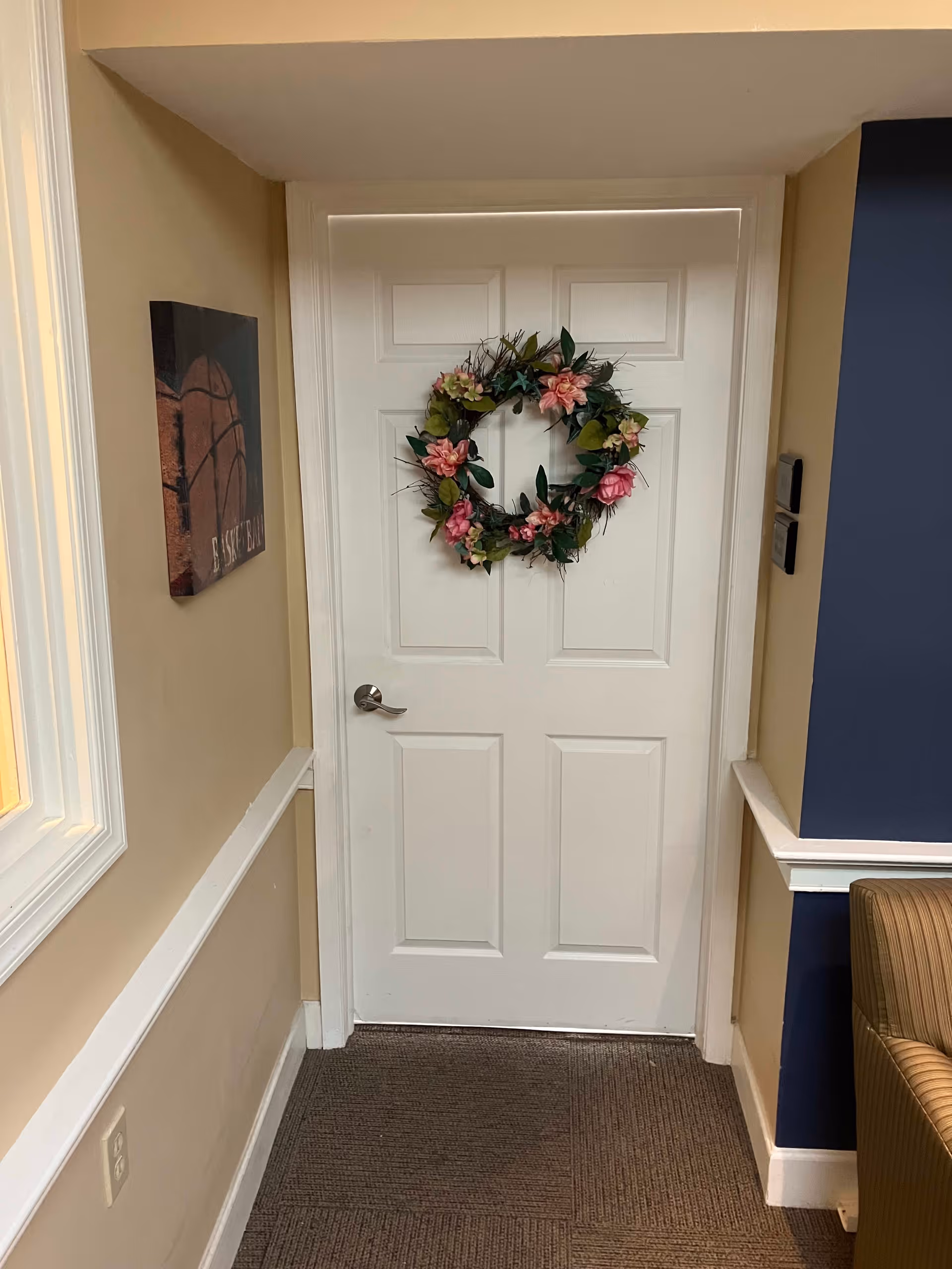 A white paneled door decorated with a floral wreath featuring pink and green flowers. The door is set in a beige and blue hallway with a brown carpeted floor. To the left of the door is a wall with a basketball-themed picture, and to the right is part of a striped armchair.