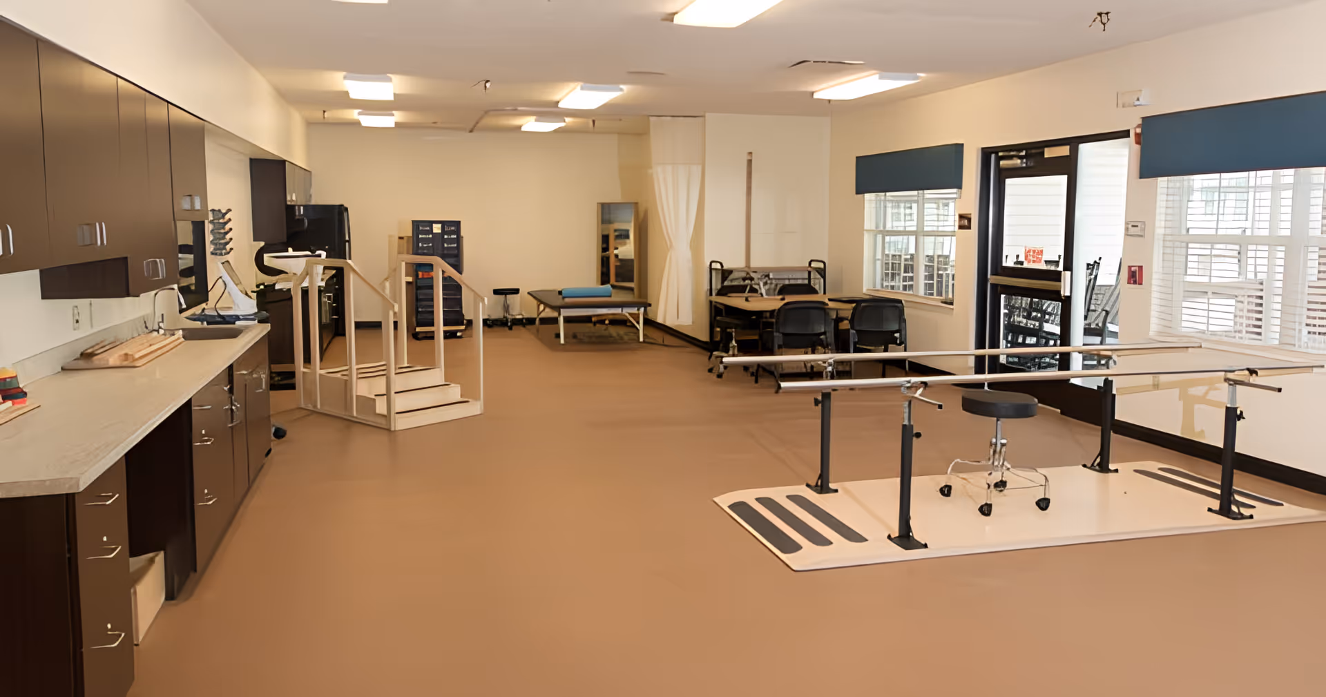 Spacious rehabilitation room with parallel bars, exercise equipment, cabinets, and seating near windows.