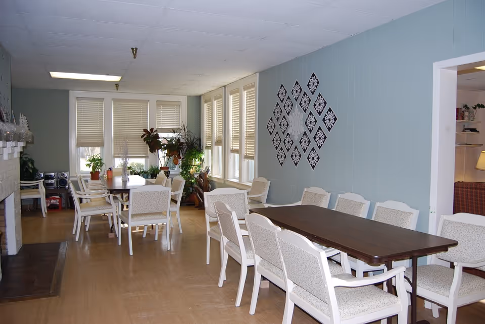 Light-filled dining room with long tables, upholstered chairs, potted plants, and decorative wall art.