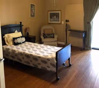 Sunlit bedroom with a single bed on castors, an upholstered armchair, a nightstand, wall art, and a window with curtains.