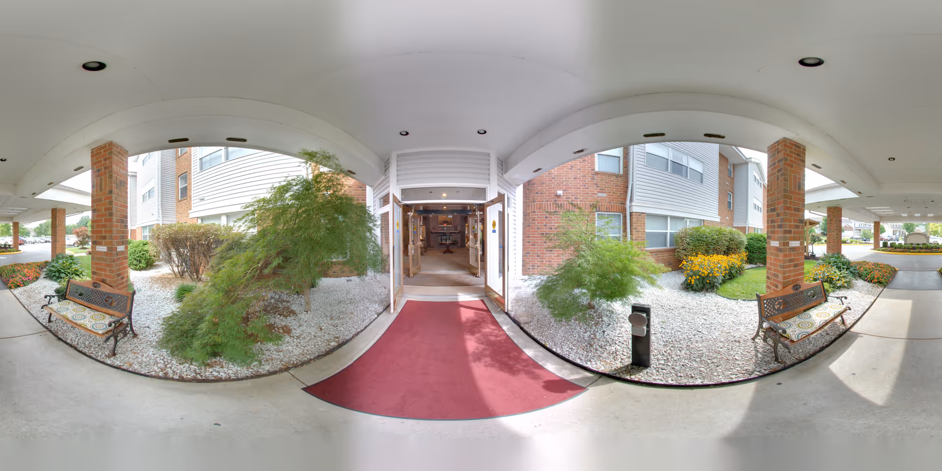 Covered entrance area of The Villa at Riverwood senior living facility with a red carpet leading to double glass doors. The entrance is flanked by brick pillars and landscaped garden beds with bushes and flowers. Two benches with patterned cushions are placed on either side of the walkway under the covered area.