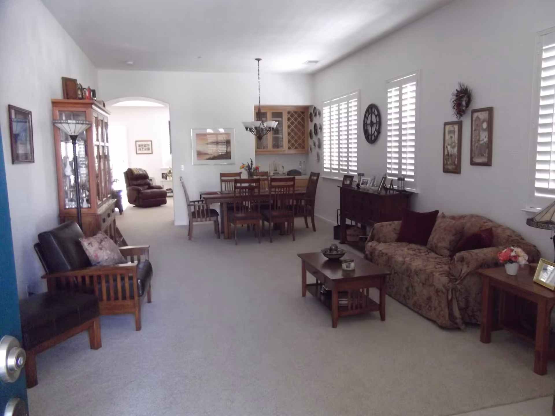 Spacious living and dining area with sofa, chairs, coffee table and dining table under a chandelier with shuttered windows.