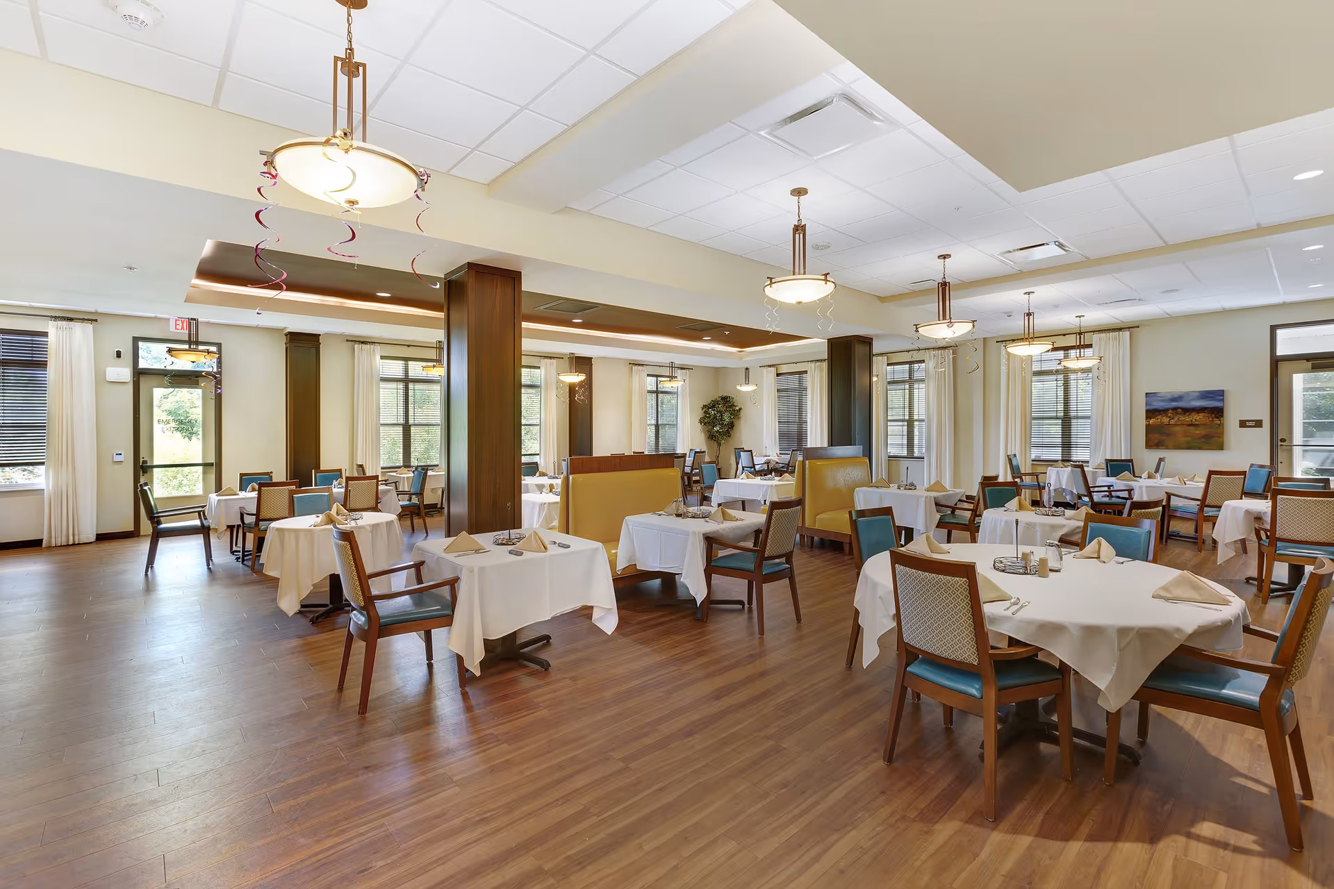 A spacious dining room with multiple tables covered in white tablecloths, each set with napkins and silverware. The room features wooden flooring, large windows with curtains, and modern hanging light fixtures. There are chairs with blue and beige upholstery and some booth seating along the walls. The room is well-lit with natural light coming through the windows.