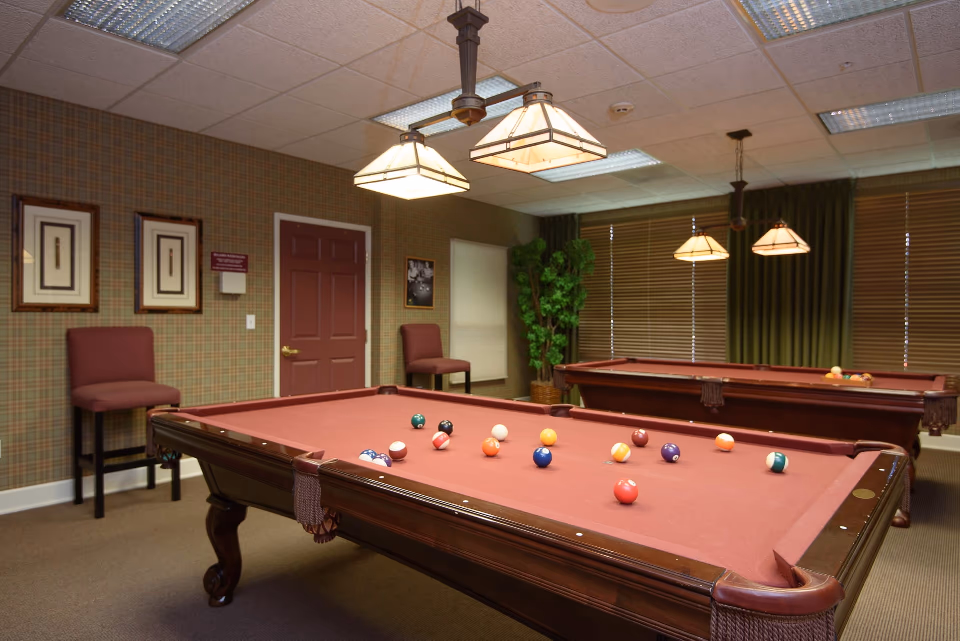 Interior room with two billiard tables covered in red felt, billiard balls scattered on the nearest table, two hanging light fixtures above each table, plaid wallpaper, two chairs against the wall, framed pictures, a potted plant, and window blinds with green curtains.