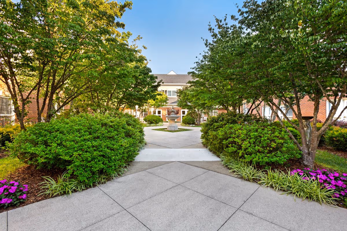 A landscaped outdoor courtyard with a concrete walkway leading to a central fountain, surrounded by green bushes, trees, and colorful flowers, with a brick building in the background under a clear blue sky.