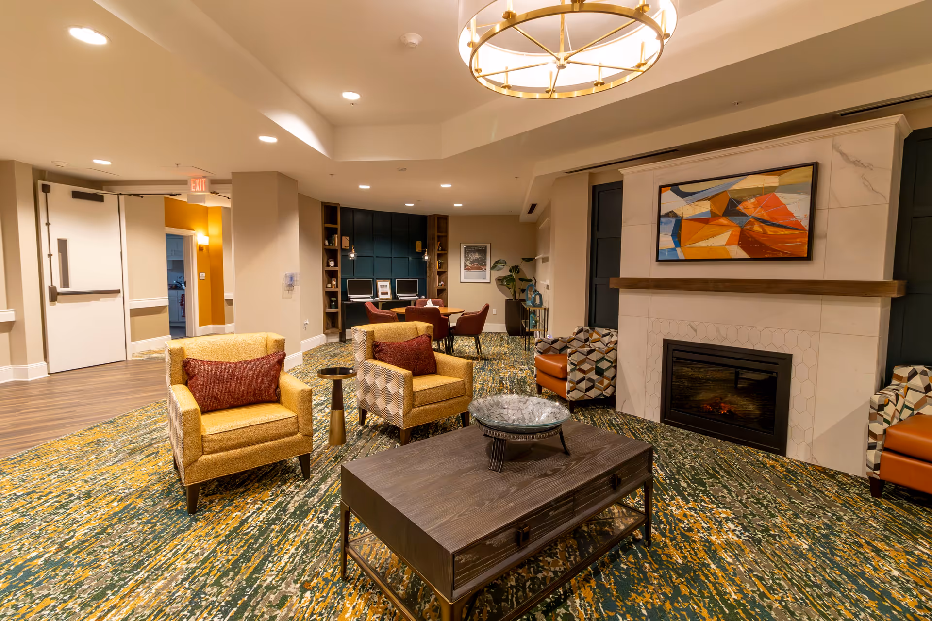 A cozy living room area in a senior living facility featuring a patterned carpet with green, yellow, and white colors. There are two yellow armchairs with red cushions, a dark wooden coffee table with a decorative bowl, and two patterned armchairs near a fireplace with a modern abstract painting above it. In the background, there is a small table with four chairs and a built-in shelving unit with two computers. The room is well-lit with recessed ceiling lights and a large circular chandelier.