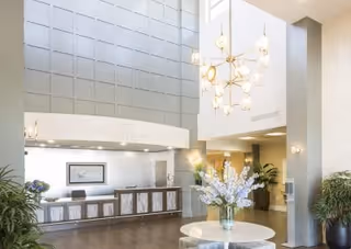 Bright two-story lobby with a reception desk, tall paneled wall, chandelier, round table with a floral arrangement, and potted plants.