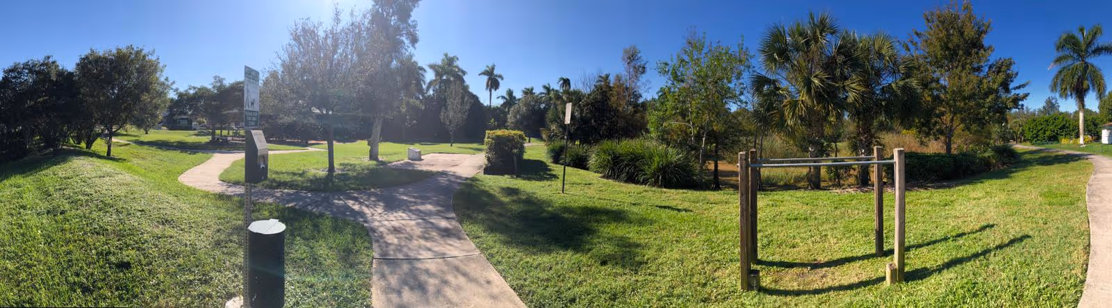 A sunny outdoor park area with green grass, palm trees, and a paved walking path that splits into two directions. There are benches, a trash can, and a small exercise or play structure visible. The sky is clear and blue.