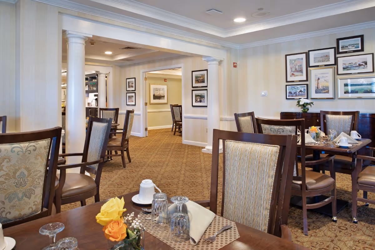 A dining room in a senior living facility with wooden tables and chairs. Tables are set with glasses, napkins, and small flower arrangements. The room has beige walls adorned with framed pictures and carpeted floors. White columns and crown molding add to the elegant decor.