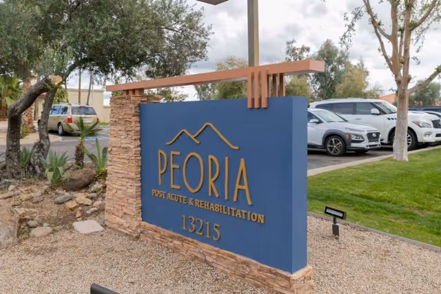 Outdoor view of a sign for Peoria Post Acute & Rehabilitation with the address 13215, surrounded by trees, plants, and parked cars in the background.