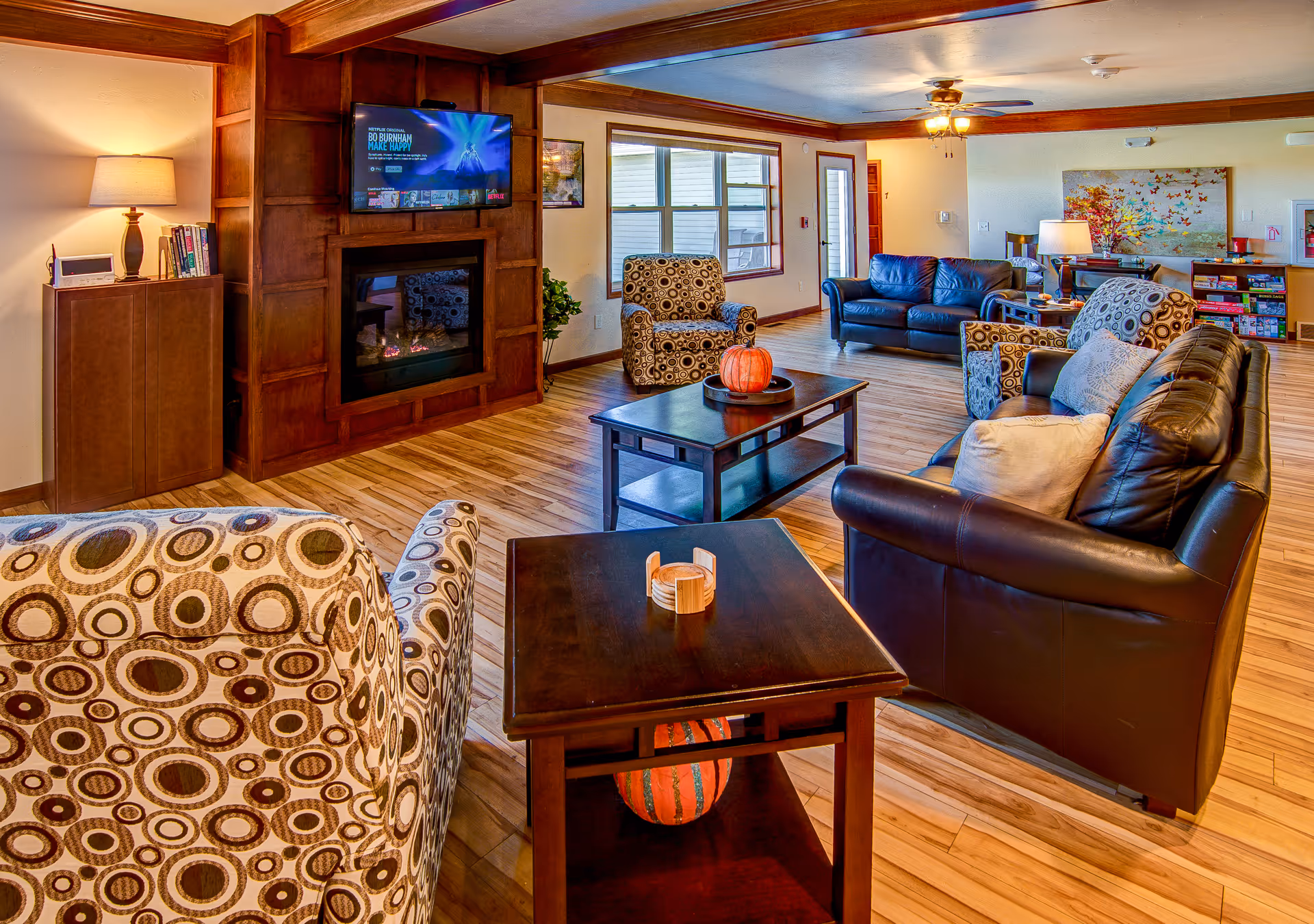 A cozy living room area in a senior living facility featuring patterned armchairs, black leather sofas, a wooden coffee table with a pumpkin decoration, a fireplace with a mounted TV above it, wooden flooring, and a ceiling fan. There is a large window and a door letting in natural light, and a colorful painting on the far wall.