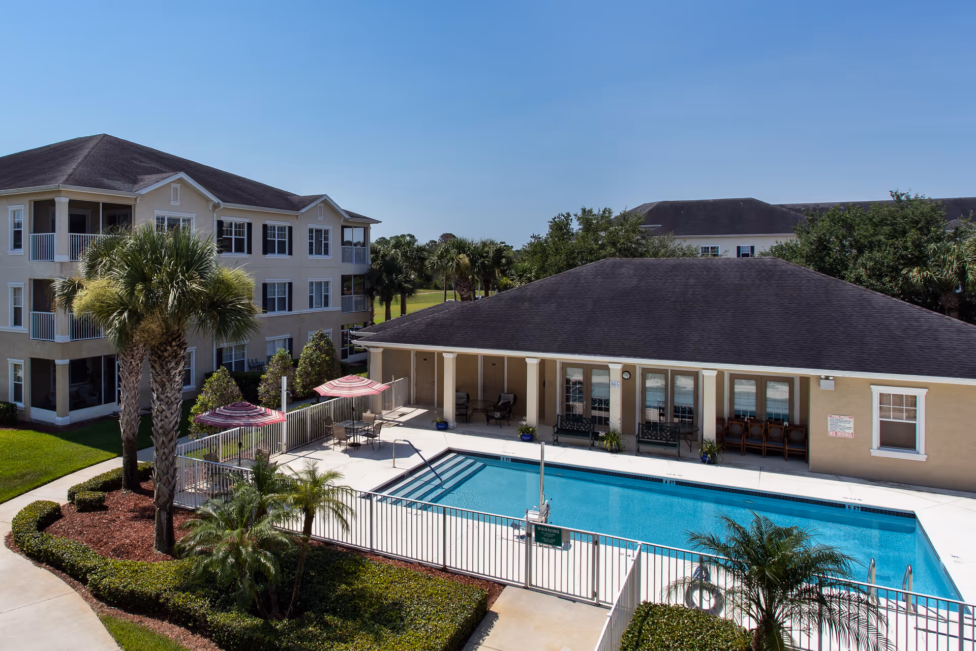 Outdoor swimming pool area at Glenbrooke Senior Living with a fenced pool, lounge chairs, tables with umbrellas, palm trees, and a beige building with multiple windows and doors in the background under a clear blue sky.