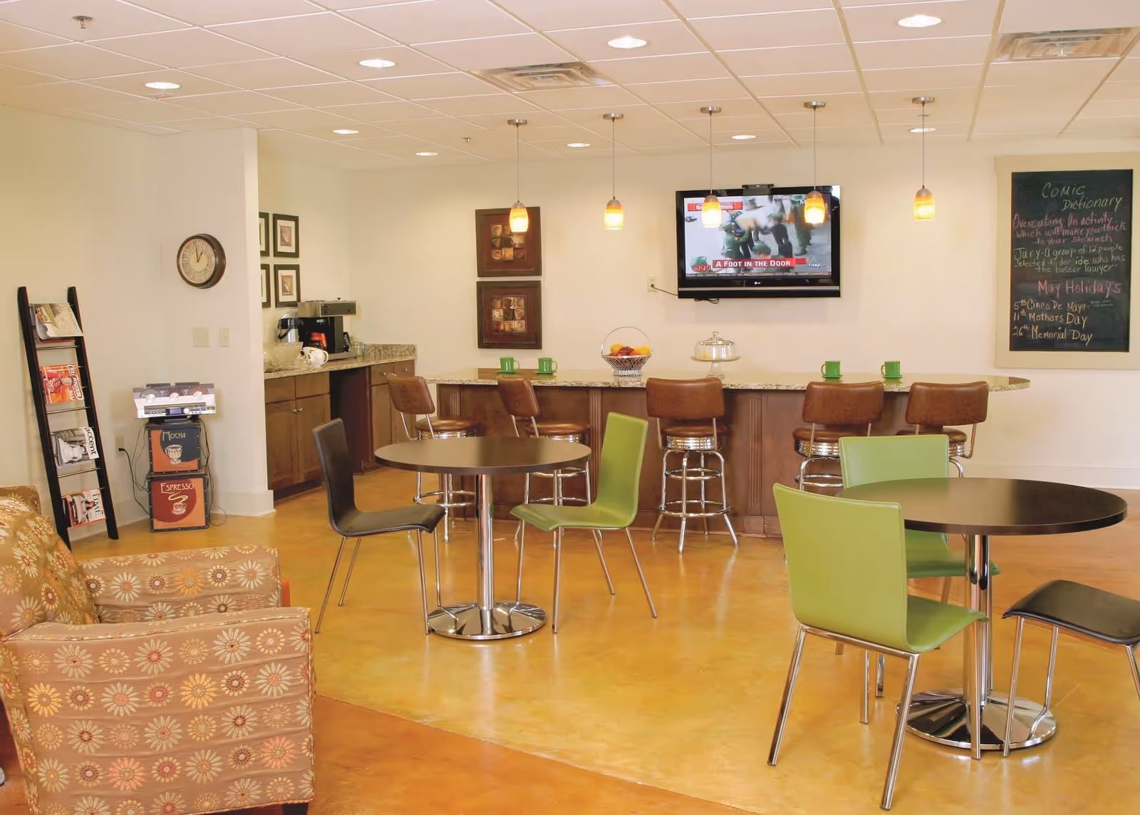 A cozy common area with a small kitchen counter and bar stools, two round tables with chairs, a patterned armchair, a wall-mounted TV showing a news channel, a coffee station, a clock on the wall, and a chalkboard with notes and holiday reminders.