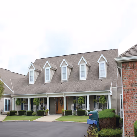 Front exterior view of a senior living facility building with a sloped roof featuring five dormer windows. The entrance has a covered porch with hanging plants and rocking chairs. There is a paved driveway and well-maintained landscaping including bushes and grass.