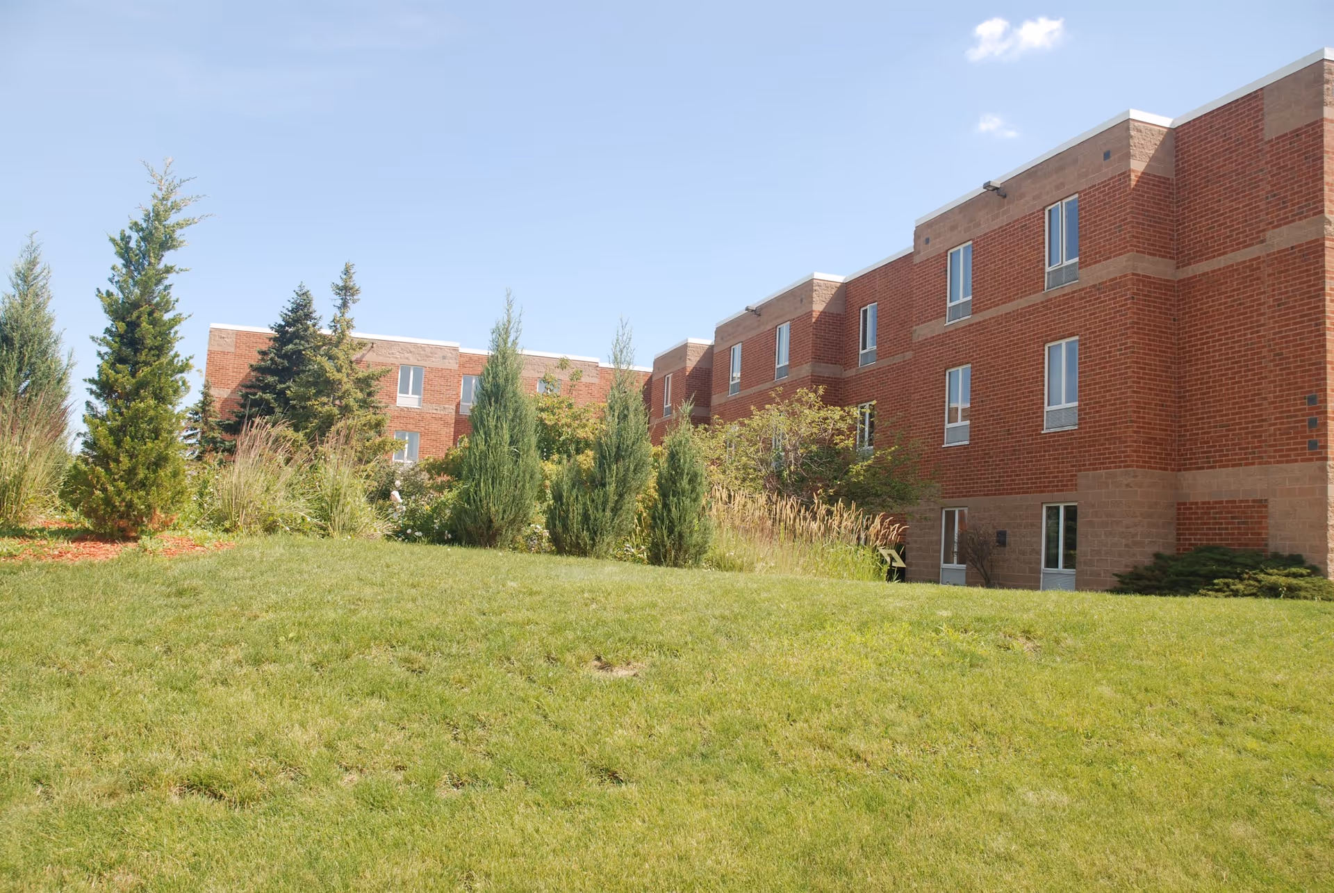 Exterior view of a multi-story red brick building with several windows, surrounded by green grass, shrubs, and small trees under a clear blue sky.