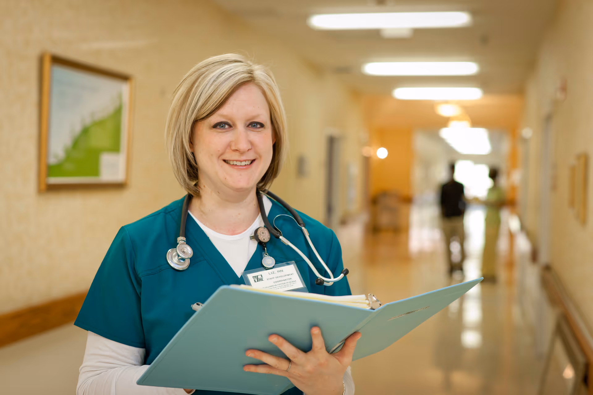A female healthcare professional wearing teal scrubs and a stethoscope around her neck is standing in a well-lit hallway holding an open blue folder and smiling at the camera. The hallway has beige walls with framed pictures and two blurred figures walking in the background.