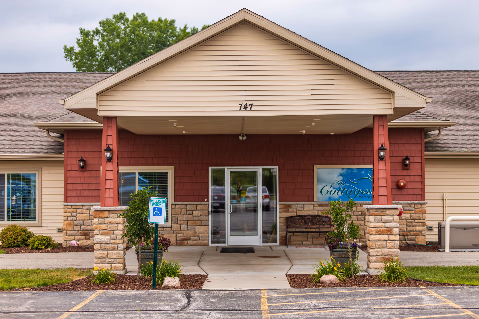Front entrance of a single-story memory care building with a covered portico, double glass doors, stone columns, landscaping, and a reserved handicap parking sign.
