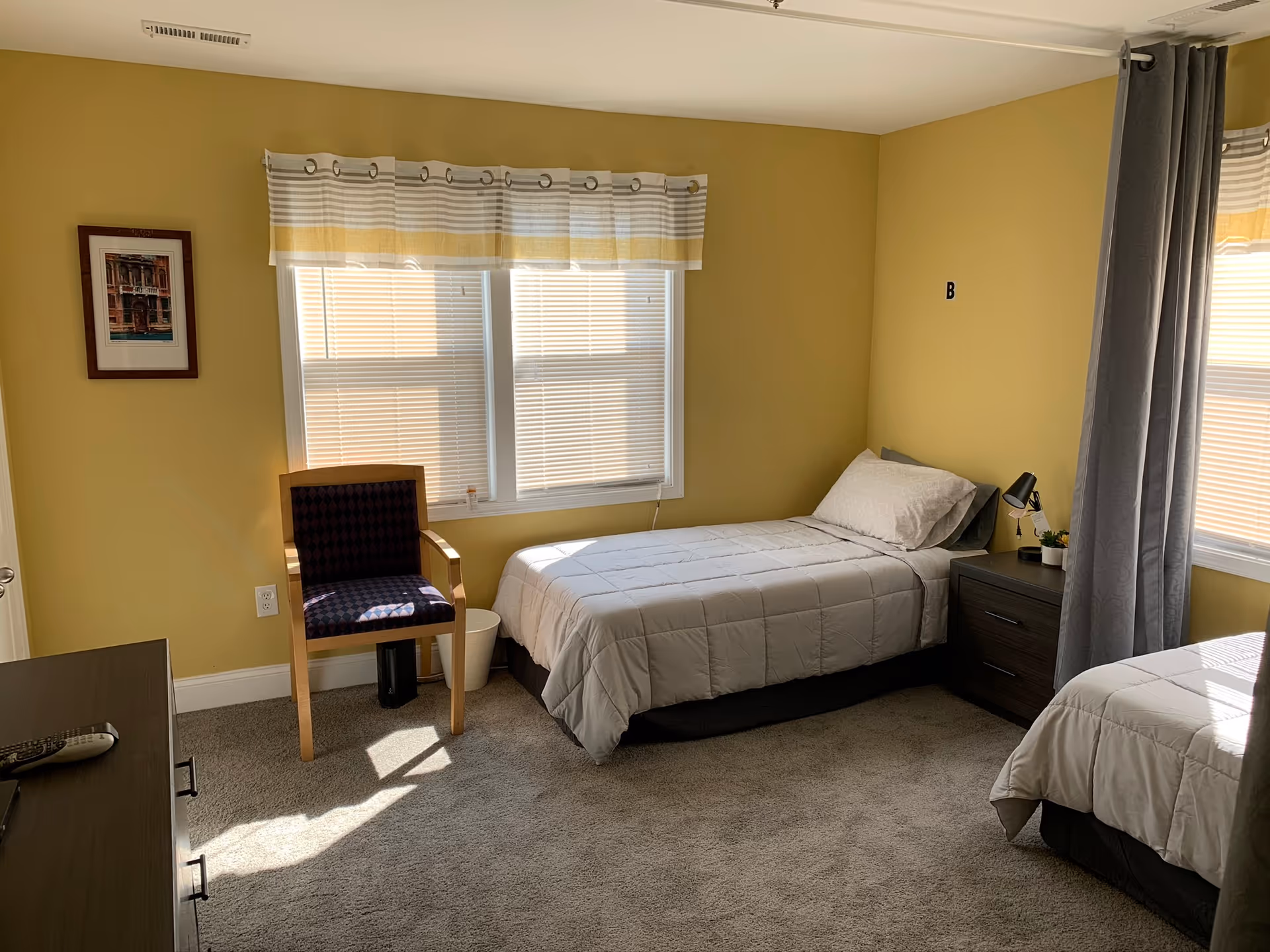 A bright bedroom with two single beds covered in light gray bedding. There is a wooden chair with a dark cushion near a window with blinds and a valance. A small nightstand with a lamp and a small plant is between the beds. The walls are painted yellow, and there is a framed picture on one wall. The carpet is gray, and sunlight streams through the windows.